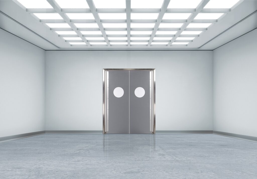 Bright, sterile room with grid ceiling lights and gray double doors with round windows.