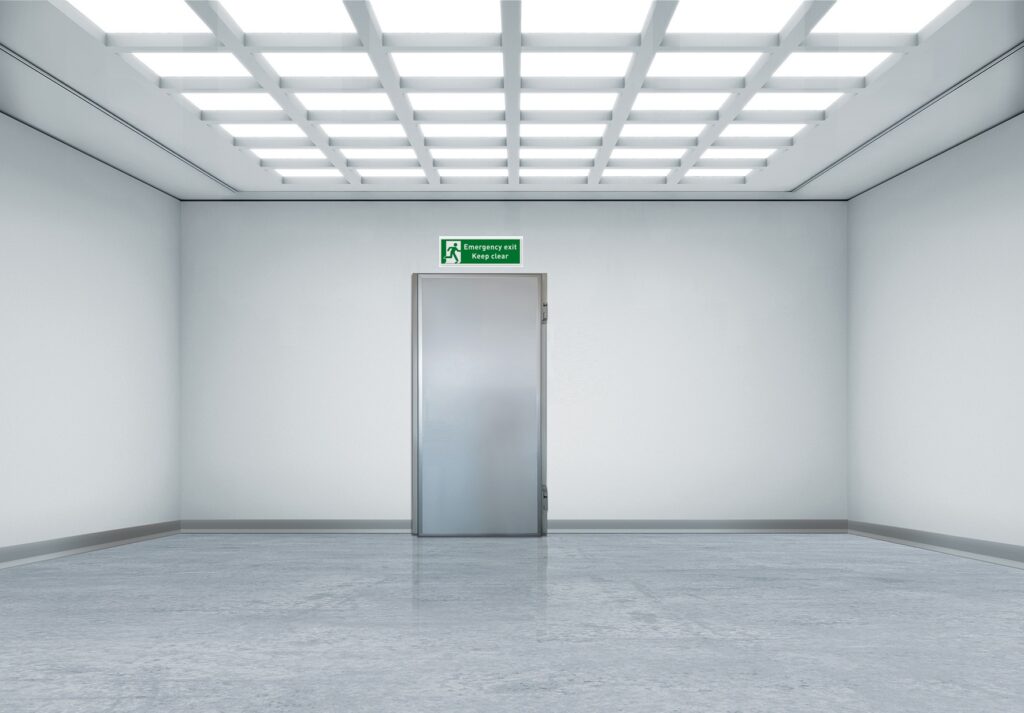 Empty white room with metal door, grid ceiling lights, and an "Emergency exit" sign.